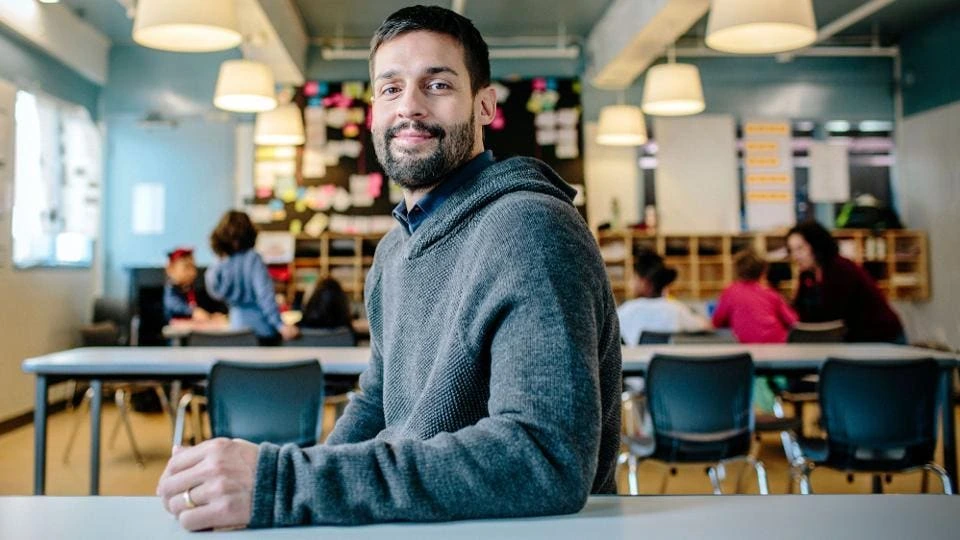 Max Ventilla seated in an AltSchool classroom, with students and classroom fixtures blurred in the background.