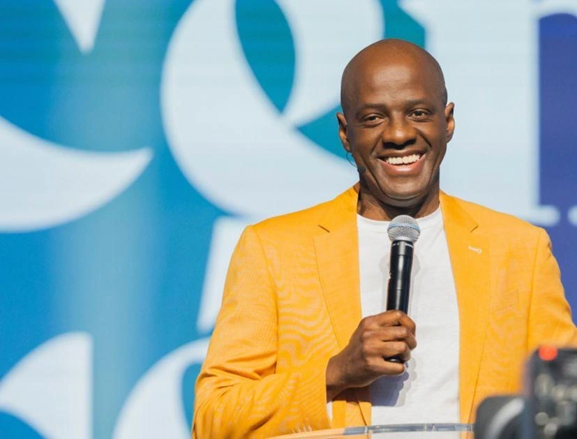Yvan Castanou smiling while speaking into a microphone on stage, wearing a bright yellow jacket.