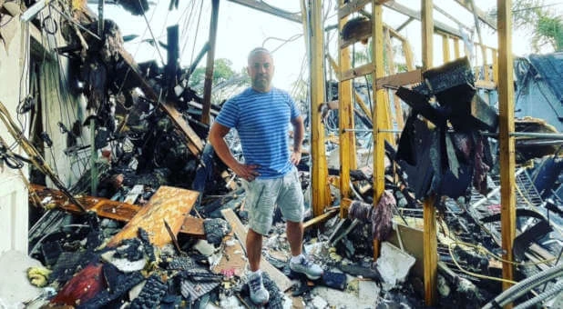 Rich Vera standing in a burned-out church interior after a fire, wearing a striped shirt and shorts